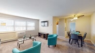 This single-floor residence offers a thoughtfully organized layout that flows naturally between its key living spaces. The living room serves as the heart of the unit, featuring wall-to-wall carpet, a wall-mounted flat-screen TV above a media console with an electric fireplace insert, and a wide window fitted with blinds that allows natural light to fill the room. A wall-unit air conditioner and baseboard heating ensure year-round comfort.

The living area transitions seamlessly into a dedicated dining space, accommodating a full dining table and chairs beneath a ceiling-mounted light fixture. Adjacent to this, the kitchen is equipped with warm-toned wood cabinetry, dark granite-style countertops, a stainless steel sink, a mosaic tile backsplash, and a window overlooking the exterior. A full-size refrigerator and dishwasher are also present.

The bedroom is a calm, carpeted retreat with neutral walls, a window providing natural light, and ample space for a queen-size bed with nightstands. The bathroom features a tiled shower-tub combination with a curtain and clean, simple fixtures. The unit's entry area provides additional open space, completing a practical and comfortable overall layout.