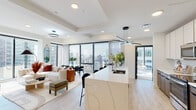 This contemporary high-rise residence offers a thoughtfully designed open-concept layout with light hardwood flooring, recessed lighting, and a bright, neutral palette throughout. The expansive living area flows seamlessly from the kitchen, anchored by floor-to-ceiling windows that frame sweeping panoramic city views and fill the space with abundant natural light.

The chef's kitchen is a standout feature, showcasing a generous waterfall-edge marble island with bar seating, sleek white upper cabinetry paired with warm wood lower cabinets, stainless steel appliances, and a striking geometric pendant light overhead. A dedicated dining area sits adjacent, offering additional city vistas.

The living room is generous and airy, centered around a wall-mounted television on a floating media console, with direct access to a private balcony overlooking the urban skyline. The primary bedroom is bathed in natural light through expansive corner windows, creating a bright and serene retreat. The en-suite bathroom features a double vanity with backlit mirror, marble-tiled walk-in shower with glass enclosure, and wood-toned cabinetry. A second bathroom offers clean, modern finishes with a tub-shower combination. Additional rooms include a flexible secondary bedroom or office space with city views, completing this sophisticated urban residence.