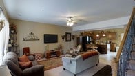 This inviting 2-floor home is designed with comfort and functionality in mind. The main level showcases an open-concept layout where the living room flows naturally into the kitchen and dining area, creating a cohesive and social atmosphere. The living room is warmly appointed with plush seating, including a light gray sofa accented with a cozy throw blanket, complemented by a patterned area rug over light carpeting. A wall-mounted flat-screen television serves as a focal point, flanked by decorative wall accents that add character to the space. A ceiling fan with integrated lighting provides both comfort and illumination. The kitchen area, visible from the living room, features warm wood cabinetry, pendant lighting, and a functional layout with ample counter space. A raised breakfast bar with seating offers a casual dining option, while the open sightlines between rooms enhance the sense of spaciousness. Warm neutral tones run throughout the main living area, punctuated by natural greenery and decorative accents that give the space a homey, personalized feel. The second floor adds additional living space, rounding out a well-proportioned home suitable for everyday living and entertaining alike.