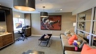 Located in Arlington, Texas, this single-floor space blends functional design with contemporary aesthetics. The main living and lounge area serves as the centerpiece, featuring plush, neutral-toned seating accented with vibrant coral and patterned throw pillows, creating a welcoming yet polished atmosphere. A sleek metal and glass coffee table anchors the seating arrangement, complementing the space's modern sensibility.

A striking large-scale abstract painting in deep reds, blacks, and whites dominates one wall, serving as a bold focal point that elevates the room's visual character. Overhead, a cylindrical drum pendant light provides warm, focused illumination, while recessed lighting throughout ensures the space remains well-lit.

A dedicated workspace is integrated seamlessly into the layout, complete with a built-in desk surface, dark cabinetry, and ergonomic office seating — making the space as practical as it is stylish. The neutral carpeted flooring and warm gray walls provide a cohesive backdrop that ties together the various functional zones. Overall, this space offers a sophisticated and versatile environment suited for both relaxation and productivity.