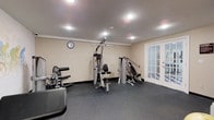 This single-level fitness room offers a dedicated space for strength and resistance training, designed with functionality in mind. The room features neutral beige walls with crown molding detail and a dark, rubber-textured flooring that provides durability and traction during workouts. Recessed ceiling lights illuminate the space evenly, creating a bright and practical environment suitable for exercise.

The room is equipped with multiple cable and weight machines, including a multi-function cable station and a dual-purpose strength machine, accommodating a variety of upper and lower body exercises. An additional piece of cardio or resistance equipment is positioned near the entrance, maximizing use of the available floor space. A wall clock is mounted prominently, allowing users to monitor their workout time with ease.

A set of elegant French doors with glass panels is located along one wall, providing access to an adjacent area and allowing natural light to filter into the room. The overall layout is organized and efficient, making the most of the available space while maintaining clear pathways between equipment.
