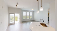 This well-appointed single-floor residence features a thoughtfully designed open-concept layout with light wood-style flooring and a neutral palette throughout. The living and dining area is bathed in natural light from a bank of windows fitted with blinds, framing expansive views of the surrounding landscape. Gold-accented pendant lights hang above the kitchen island, complementing the stainless steel sink set into white quartz countertops. The kitchen is fully equipped with grey cabinetry, a full suite of stainless steel appliances—including a refrigerator, range, built-in microwave, and dishwasher—and a sleek tile backsplash. A track lighting fixture illuminates the space effectively. A private balcony is accessible through glass-paned doors from the living area. The bedrooms are generously proportioned with clean white walls and include access to a spacious walk-in closet with multi-tiered shelving and hanging rods. The primary bathroom features a double-sink vanity with a wide mirror, a soaking tub, and tiled shower surround, while the secondary bathroom includes a tub-shower combination and wall-mounted storage cabinet.