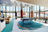 This expansive single-floor amenity space is thoughtfully designed to serve as a dynamic social hub, combining recreation, dining, and relaxation within one cohesive layout. The centerpiece of the main lounge is a sleek pool table anchored by a striking, organically shaped teal area rug with coral and earth-tone borders, adding artistic flair to the generous open floor plan.

Soaring double-height ceilings in the primary lounge area are adorned with a bold, large-scale artistic mural, complemented by floor-to-ceiling glass walls that flood the space with natural light and frame panoramic urban views. Terracotta-hued concrete columns punctuate the room, adding an industrial warmth to the modern aesthetic.

A dedicated lounge zone features built-in shelving, a media wall, and comfortable seating beneath a warm wood-paneled ceiling. The kitchen area offers light blue cabinetry, a textured geometric tile backsplash, a marble-topped island with bar seating, draft taps, and patterned cement tile flooring.

Sliding glass doors open to a generous outdoor terrace furnished with wraparound upholstered seating and a fire pit, offering open sky views of the surrounding cityscape, seamlessly extending the indoor experience outdoors.