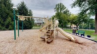Located in Baltimore, MD, this outdoor tot lot is a thoughtfully designed recreational space that offers a welcoming environment for children and families. At the heart of the space is a multi-component playground structure finished in neutral tan tones, featuring multiple slides, a climbing ladder with rope elements, monkey bars, staircase access points, and elevated walkways with safety railings. The playground is set on a generous wood chip surface that provides cushioning and defines the play zone clearly within the surrounding landscape.

The grounds are well-kept, with neatly manicured green lawns extending outward from the play area. Mature deciduous and evergreen trees provide natural shade and a lush canopy throughout the space, contributing to a calm and pleasant atmosphere. A black wrought iron perimeter fence encloses part of the lot, offering a secure boundary while maintaining open sightlines to the surrounding neighborhood.

Adjacent to the play area, a paved seating pad with a green bench and a waste receptacle provides a convenient resting spot for caregivers and visitors. A nearby brick residential building with classic multi-pane windows adds architectural context to the setting. Informational signage is posted at the entrance. The overall ambiance is clean, safe, and community-oriented, making this tot lot a functional and inviting outdoor amenity.