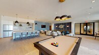 This contemporary amenity lounge is a well-appointed, open-plan space designed to accommodate a variety of social and recreational activities. Light wood-look tile flooring flows throughout, lending a clean, cohesive aesthetic that complements the neutral color palette of soft whites, warm creams, and calming blue-gray accent walls.

The space is organized into distinct functional zones. A game area anchors one side of the room, featuring a full-size pool table beneath a striking multi-shade pendant light fixture with gold interior accents. A wall-mounted flat-screen TV is positioned nearby, adding to the entertainment options. Adjacent to this is a cozy lounge zone with plush upholstered sofas and armchairs arranged around low coffee tables, ideal for casual conversation or relaxation. A decorative textured wall panel with sculptural accents and flanking wall sconces adds an artistic focal point to this seating area.

The kitchenette bar is outfitted with a large stainless steel refrigerator, an iridescent tile backsplash, open shelving with greenery, a sink, and a spacious counter with bar seating — perfect for hosting. A high-top table positioned beside a large window offers views of the surrounding outdoor greenery, filling the corner with natural light. Recessed ceiling lighting throughout ensures the entire space remains bright and welcoming at all hours.