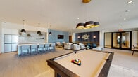 This contemporary amenity lounge is a well-appointed, open-plan space designed to accommodate a variety of social and recreational activities. Light wood-look tile flooring flows throughout, lending a clean, cohesive aesthetic that complements the neutral color palette of soft whites, warm creams, and calming blue-gray accent walls.

The space is organized into distinct functional zones. A game area anchors one side of the room, featuring a full-size pool table beneath a striking multi-shade pendant light fixture with gold interior accents. A wall-mounted flat-screen TV is positioned nearby, adding to the entertainment options. Adjacent to this is a cozy lounge zone with plush upholstered sofas and armchairs arranged around low coffee tables, ideal for casual conversation or relaxation. A decorative textured wall panel with sculptural accents and flanking wall sconces adds an artistic focal point to this seating area.

The kitchenette bar is outfitted with a large stainless steel refrigerator, an iridescent tile backsplash, open shelving with greenery, a sink, and a spacious counter with bar seating — perfect for hosting. A high-top table positioned beside a large window offers views of the surrounding outdoor greenery, filling the corner with natural light. Recessed ceiling lighting throughout ensures the entire space remains bright and welcoming at all hours.