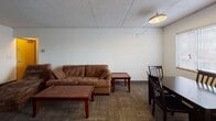 This single-floor unit offers a straightforward, functional layout designed to accommodate everyday living. The living area provides a comfortable gathering space with carpeted flooring and a suspended pendant light fixture overhead, complemented by a drop tile ceiling that is consistent throughout the unit. A dedicated dining area adjacent to the living space features a window fitted with horizontal blinds, allowing natural light to filter in, and offers enough room for a full dining table set.

The kitchen is compact yet efficiently arranged, featuring an electric coil range, overhead exhaust hood, and ample upper cabinetry with wood-trimmed accents. A sink and additional counter space complete the workspace, with tile flooring providing a practical surface underfoot.

The unit includes two bedrooms, each carpeted and furnished with a window that brings in natural light. One bedroom includes a built-in desk unit with shelving and storage drawers, making it well-suited for study or work. The bathroom is equipped with a wood-toned vanity with ample drawer storage, a large mirror, and notable accessibility features including multiple grab bars positioned near the toilet area, alongside vinyl flooring throughout.