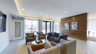 Located at 3443 Park Street in Grove City, Ohio, this single-floor space offers a thoughtfully designed interior that blends modern aesthetics with functional comfort. The lounge area serves as a welcoming centerpiece, outfitted with a curated mix of contemporary seating including a tufted dark sofa accented with teal throw pillows, upholstered armchairs, and a rounded leather ottoman that anchors the arrangement. Recessed lighting is complemented by warm cove lighting along the ceiling edge, casting a soft ambient glow throughout the space. A striking natural wood accent wall adds warmth and texture, adorned with framed abstract artwork that contributes to the space's modern character. Floor-to-ceiling windows and glass entry doors along the front facade flood the interior with abundant natural light while offering clear views of the surrounding outdoor environment. Sheer white drapery panels frame the windows, softening the bright exterior light. The light-toned flooring unifies the open layout, while clean white walls and crisp architectural lines maintain a polished, contemporary feel. Overall, the space conveys an inviting and professional ambiance well-suited for community gathering or reception use.