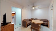 This single-floor space is designed with practicality in mind, combining a living and dining area within one open room. The living area is furnished with a modest sofa and matching armchairs in a neutral brown tone, arranged to encourage casual conversation or relaxation. An octagonal wooden dining table with coordinating chairs sits nearby, making efficient use of the shared space. Overhead, a ceiling fan with integrated lighting provides both ventilation and illumination, contributing to a comfortable atmosphere. The walls are finished in clean white, and the light-toned tile flooring runs throughout, lending a bright, uniform feel to the interior. A doorway connects to a separate kitchen, which is visible through the opening and features warm wood cabinetry, white appliances including a refrigerator and dishwasher, and its own tile flooring. The kitchen is compact but functional, providing all essential cooking amenities. Overall, the space maintains a simple, no-frills aesthetic, suited for everyday living with a focus on utility and ease of use.
