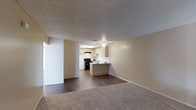 This single-floor space offers an open-concept layout that connects the living area to the kitchen, creating a cohesive and functional floor plan. The living area features neutral-toned carpet, while the kitchen and transitional zones showcase dark hardwood-style flooring, providing a clear visual distinction between spaces. Walls are painted in a warm beige tone throughout, contributing to a consistent and understated aesthetic.

The kitchen is positioned at the rear of the main living space and is equipped with a black gas range, a range hood, and light wood-toned cabinetry that lines both the upper and lower walls. A peninsula counter with matching cabinetry serves as a natural divider between the kitchen and the living area, offering additional workspace and a casual breakfast bar setup. A skylight above the kitchen brings in natural light, brightening the cooking area. A ceiling fan is centrally mounted in the kitchen zone, aiding in air circulation. The textured popcorn ceiling is consistent throughout the space, and large baseboards frame the perimeter of each room. The overall ambiance is practical and move-in ready.