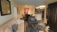This single-floor residence is designed around an open-concept floor plan that seamlessly connects the living, dining, and kitchen areas into one cohesive, functional space. The living area serves as the central gathering point, furnished with a light-toned sofa and a patterned accent chair arranged around a glass-top coffee table, creating a comfortable and inviting atmosphere. Warm yellow-beige walls, accented by crown molding, lend a classic touch to the interior, while the dark hardwood-style flooring provides a rich contrast throughout.

Adjacent to the living area, the dining space accommodates a round table with seating for several guests, lit by a table lamp that adds a soft, warm glow. A bold abstract artwork on the wall serves as a focal point for this zone. The kitchen is positioned just beyond, featuring dark cabinetry, a breakfast bar with a raised counter, stainless steel appliances, and tile flooring, distinguishing it subtly from the rest of the space. Upper cabinet surfaces are adorned with decorative greenery, adding a lived-in charm. The overall ambiance is warm, cohesive, and well-appointed.