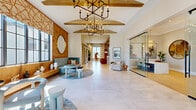 Located at 11590 SW Roma Way in Port Saint Lucie, Florida, this single-story community clubhouse delivers a sophisticated and welcoming atmosphere through a carefully curated blend of contemporary design elements. The expansive entry foyer greets visitors with large-format porcelain tile flooring, soaring vaulted ceilings with white shiplap detailing, and exposed wood beam accents. A striking chandelier anchors the grand entry hall, while floor-to-ceiling glass doors frame views of the surrounding palm-lined exterior. The main lounge area features a distinctive textured scallop-patterned accent wall in warm amber tones, complemented by a curved powder-blue seating arrangement and abundant natural light from oversized black-framed windows. An open-concept recreation room showcases light wide-plank hardwood flooring, woven rattan pendant lighting, and dual pool tables, creating a lively yet refined social space. An adjacent wet bar and lounge area offers a long quartz-topped counter with stylish seating. A glass-enclosed private office suite provides a sleek workspace, while a secondary lounge area features built-in open shelving, a white fireplace surround, and curated décor. The overall ambiance is bright, modern, and resort-inspired, reflecting the upscale character of the Port Saint Lucie community it serves.