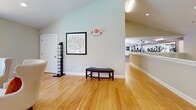 Located at 3250 Mercer University Dr in Chamblee, GA, this two-floor commercial space offers a thoughtfully designed layout spanning a generous total area across both levels. The ground floor houses a well-equipped fitness center with rubber flooring, a multi-station cable machine, a stationary bike, treadmill, and a wall-mounted TV — all benefiting from natural light through large windows overlooking greenery outside. Two ADA-accessible bathrooms with grab bars and tile flooring serve the space conveniently. A welcoming hallway with vaulted ceilings and warm hardwood flooring connects areas seamlessly. The kitchen features crisp white shaker cabinetry, dark granite countertops, a teal accent wall, and stainless steel appliances, providing a fully functional prep area. The upper floor expands the space considerably, highlighted by a stylish living room with soaring ceilings, a white fireplace framed by deep green accent walls, a statement chandelier, and hardwood floors. A formal conference/dining room accommodates a large walnut table with black leather seating and a wall-mounted display, ideal for meetings or gatherings. An additional private office with a pass-through window completes the upper level, creating a versatile, multi-purpose environment throughout.