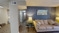 This single-story residence offers a well-organized layout with distinct living, dining, sleeping, and kitchen areas. The living room serves as the focal point of the home, featuring durable wood-look flooring, a bold dark blue accent wall, and a large sliding glass door fitted with vertical blinds that allows natural light to fill the space. A hallway connects the main living area to the rest of the unit.

The kitchen is designed in a galley-style configuration with ample white cabinetry, light countertops, a double-basin sink, and white appliances including a refrigerator, range, and dishwasher. Wood-look flooring continues throughout this space. Adjacent to the kitchen, the dedicated dining area accommodates a round glass-top table beneath a ceiling fan with integrated lighting, and features the same wood-look flooring.

The unit includes two bedrooms, both carpeted and finished with neutral gray walls and textured ceilings. The primary bedroom offers natural light from a window with sheer curtains, while the secondary bedroom is comparably sized and similarly appointed. The bathroom features tiled wainscoting, dark upper walls, a vanity with a white countertop, and a tub-shower combination.