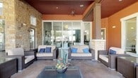 This single-story space showcases a sophisticated contemporary design with seamless transitions between indoor and outdoor living areas. The covered outdoor patio is anchored by a stone fireplace and features a wood-paneled ceiling with a ceiling fan, comfortable wicker seating with neutral cushions and blue accent pillows, and large sliding glass doors that connect directly to the interior. Natural stone columns frame the patio, adding a warm, textural contrast to the modern design.

Inside, the living area features a striking floor-to-ceiling textured tile accent wall with a wall-mounted television, complemented by a starburst chandelier and a rustic wood console table with metallic accents. Large sliding glass doors flood the space with natural light and maintain visual continuity with the outdoor area.

The kitchen is a standout, featuring a substantial stainless steel island with sleek black and chrome bar seating, geometric diamond-shaped pendant lights, a shimmering mosaic tile backsplash, and rich walnut cabinetry paired with a panel-ready refrigerator. A full-glass interior door connects the kitchen to adjacent spaces.

The expansive game room boasts soaring ceilings, large windows, a bold navy accent wall, tile flooring, and a full-size pool table as its centerpiece.