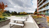 This expansive outdoor common area is thoughtfully designed to offer a variety of recreational and social amenities. The grounds feature wide, aggregate concrete pathways that wind through the space, providing accessible circulation between distinct zones. A standout feature is the putting green, offering a well-maintained recreational outlet set against a backdrop of mature deciduous trees displaying rich autumn foliage.

Multiple picnic tables are arranged across an open patio area, creating a comfortable setting for outdoor gatherings. Stone-edged raised planters and garden beds are integrated throughout, filled with an array of ornamental grasses, shrubs, and seasonal plantings in warm hues of red, orange, and gold, contributing vibrant color and natural texture to the environment.

The landscaping transitions seamlessly between the patio, garden areas, and open lawn spaces, creating a layered and visually engaging outdoor environment. The multi-story red brick building flanking the space features numerous private balconies overlooking the grounds below, reinforcing the connection between interior and exterior living. The surrounding tree canopy and neighboring low-rise structures provide a sense of enclosure and community, while the open sky above contributes to the area's spacious, park-like atmosphere.