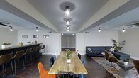 This single-floor space is thoughtfully designed to serve as a versatile communal gathering area. At its center sits a long, solid wood communal dining table surrounded by an eclectic mix of colorful chairs — including vibrant orange and blue seats — creating a lively yet welcoming atmosphere. A row of industrial-style pendant lights runs along the ceiling above the table, providing focused illumination while adding a contemporary design touch.

To one side, a bar-height counter with bar stools offers an additional seating option, ideal for casual interaction or individual use. On the opposite side, a comfortable lounge area features a dark gray sectional sofa paired with a patterned area rug, offering a more relaxed setting within the same open layout.

The space is finished with dark hardwood flooring that contrasts cleanly against the light gray walls and ceiling. Flush-mount ceiling lights and recessed fixtures ensure the entire room is well-lit. The open-plan layout encourages flow and connection between the dining, bar, and lounge zones, making it a functional and inviting environment suited for group gatherings, meetings, or everyday social interaction.