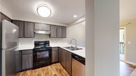 This single-floor residence offers a clean, contemporary design with a cohesive neutral color palette throughout. The kitchen is a standout feature, outfitted with dark wood-tone cabinetry, crisp white countertops, and light hardwood-style flooring. A full suite of black and stainless steel appliances — including an electric range, dishwasher, and refrigerator — provides excellent functionality. The kitchen is configured in an L-shape with a peninsula that includes a double stainless steel sink, offering an open connection to the adjacent living area. Recessed lighting and a flush-mount ceiling fixture keep the space well-lit. The bathroom is finished with a dark wood vanity topped with a white countertop and an integrated sink, a large mirror with overhead lighting, and a full tub-shower combination with chrome fixtures. Light hardwood-style flooring extends into the bathroom, complementing the warm, neutral tones of the walls. The bedroom and living areas feature wall-to-wall carpet in a soft beige tone, creating a comfortable and quiet atmosphere. Large windows in these rooms allow natural light to fill the space, with views of lush greenery outside. The overall layout is efficient and well-connected, offering a comfortable and modern living environment.