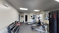 This fitness room is designed to accommodate a well-rounded workout experience within a compact, efficiently organized space. The room features vaulted ceilings fitted with two large skylight panels that flood the interior with bright, even artificial lighting, contributing to an open and energizing atmosphere. The walls are finished in a clean, neutral off-white tone, complemented by gray rubberized flooring ideal for high-impact exercise and equipment placement. A dumbbell rack lines one wall, stocked with a comprehensive range of free weights, alongside a weight plate. Cardio equipment includes a treadmill and an elliptical machine, both centrally positioned for ease of use. A cable resistance machine is also present, offering versatile strength training options. A full-length mirror panel is mounted along one wall, enhancing the sense of space and allowing users to monitor their form. Natural light enters through a window on one side of the room, providing a pleasant connection to the outdoors. Two doors provide access points, and a notice board is mounted on the wall, likely displaying fitness guidelines or facility rules.