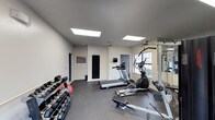 This fitness room is designed to accommodate a well-rounded workout experience within a compact, efficiently organized space. The room features vaulted ceilings fitted with two large skylight panels that flood the interior with bright, even artificial lighting, contributing to an open and energizing atmosphere. The walls are finished in a clean, neutral off-white tone, complemented by gray rubberized flooring ideal for high-impact exercise and equipment placement. A dumbbell rack lines one wall, stocked with a comprehensive range of free weights, alongside a weight plate. Cardio equipment includes a treadmill and an elliptical machine, both centrally positioned for ease of use. A cable resistance machine is also present, offering versatile strength training options. A full-length mirror panel is mounted along one wall, enhancing the sense of space and allowing users to monitor their form. Natural light enters through a window on one side of the room, providing a pleasant connection to the outdoors. Two doors provide access points, and a notice board is mounted on the wall, likely displaying fitness guidelines or facility rules.