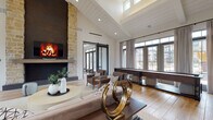 This single-story space showcases a modern farmhouse aesthetic defined by dramatic vaulted ceilings clad in white shiplap, creating an airy, expansive atmosphere throughout. The open-concept layout flows seamlessly between the main living area, a dedicated lounge zone, and a central kitchen, all unified by light-toned hardwood-style flooring. A floor-to-ceiling natural stone fireplace column serves as the visual anchor of the living area, framing a dark-surround hearth and wall-mounted screen. Opposite, a full shuffleboard table adds a recreational focal point beneath the towering windows. The kitchen features a generously sized white island with bar seating, matte black fixtures, and bright cabinetry, all bathed in natural light from an array of large multi-pane windows. A clerestory window system above the central living space draws additional daylight deep into the interior. Sliding barn doors with dark steel frames provide flexible room separation while reinforcing the industrial-farmhouse design language. Recessed lighting and statement pendant fixtures complement the natural illumination. The overall ambiance is warm, sophisticated, and welcoming, balancing refined finishes with relaxed, communal-oriented functionality.