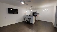 This single-floor space showcases a clean, modern design centered around an open-concept layout that seamlessly connects the living and kitchen areas. The kitchen is outfitted with crisp white cabinetry, a mosaic tile backsplash, stainless steel appliances including a refrigerator, microwave, and range, as well as recessed lighting complemented by stylish pendant lights that hang over a central breakfast bar. The breakfast bar offers casual seating with two bar stools, making it an ideal spot for dining or entertaining. The living area is anchored by rich, dark hardwood flooring that runs throughout the space, creating a striking contrast against the bright white walls. A wall-mounted flat-screen television and a ceiling fan with an integrated light fixture add both functionality and a contemporary touch to the living space. The layout is thoughtfully designed to maximize openness and flow, with soft recessed and accent lighting contributing to a warm, inviting ambiance. The minimalist aesthetic, defined by neutral tones and modern fixtures, gives the space a fresh and well-appointed feel that is both practical and visually appealing.