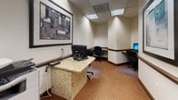 Located at 7620 Old Georgetown Road in Bethesda, Maryland, this business center offers a fully equipped, single-floor workspace designed to meet professional productivity needs. The room features a drop ceiling with recessed and panel lighting that provides consistent, even illumination throughout the space. Warm neutral walls are accented by rich wood-toned baseboards and chair rails, lending the space a polished, professional appearance. Patterned carpeting in earthy tones adds comfort underfoot while complementing the overall décor. Multiple granite-topped workstations are arranged throughout the room, each equipped with desktop computers, keyboards, and mice, offering individual semi-private work areas. A dedicated alcove workstation provides an additional secluded space for focused tasks. A multifunction printer and copier is conveniently accessible within the room, supporting a range of document needs. Framed abstract artwork adorns the walls, adding visual interest and a modern touch to the professional environment. The space is accessed through a solid door with a windowed panel, ensuring both privacy and visibility. The layout is thoughtfully organized to accommodate multiple users simultaneously while maintaining a quiet, functional atmosphere suited for business tasks.