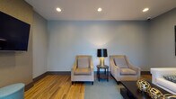 This single-floor space presents a thoughtfully designed interior that blends contemporary aesthetics with comfort and functionality. The main lounge area features rich hardwood flooring that transitions seamlessly into plush carpeting, creating distinct zones within the open layout. Soft, neutral-toned walls in muted blue-gray hues provide a calming backdrop throughout the space.

The seating area is anchored by a clean-lined white sofa and complemented by a pair of upholstered accent chairs in warm taupe, arranged around a dark wood coffee table adorned with sculptural gold décor pieces. A stylish floor lamp with a black shade and gold base adds a refined touch, while a pair of large abstract artworks in navy and gold tones bring visual interest to the walls.

A standout feature of the space is the floor-to-ceiling black-framed glass windows and doors, which flood the interior with abundant natural light and offer open views of a lush outdoor courtyard area. This glazed entry zone, finished with deep-toned hardwood flooring, creates a striking and welcoming transition into the space.

Recessed ceiling lighting provides consistent, warm illumination throughout, enhancing the modern and polished character of the interior. Overall, the space exudes a sophisticated yet approachable atmosphere, well-suited for lounging or casual gathering.