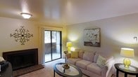 This single-floor residence offers a thoughtfully designed living area that balances comfort and functionality. The living room serves as the central gathering space, featuring a fireplace along one wall that adds both warmth and a focal point to the room. Neutral-toned walls and light carpet throughout create a soft, welcoming atmosphere that complements the understated décor.

A sliding glass door along one wall floods the space with natural light while providing direct access to an outdoor area, offering a seamless connection between interior and exterior living. Sheer curtains frame the door, allowing light to filter through while maintaining a sense of privacy.

The room accommodates a comfortable seating arrangement centered around an oval glass-top coffee table, making it well-suited for relaxing or entertaining. A wall-mounted piece of artwork featuring white florals adds a decorative accent above the sofa area. The space is enhanced by a side table with a yellow lamp, introducing a pop of color that complements the chevron-patterned accent pillows. Overall, the living area presents a clean, livable aesthetic with practical design elements throughout.