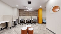 This thoughtfully designed workspace offers a dynamic blend of focused work areas and collaborative zones within a single, efficiently organized floor. The space is anchored by a row of semi-private workstations, each framed by tall acoustic divider panels that provide concentration and privacy. These stations are set against a striking textured stone accent wall, adding warmth and visual depth to the room. A bold orange structural column punctuates the interior, serving as a vibrant design focal point that ties together the space's energetic color palette.

Overhead, a distinctive X-shaped linear pendant light fixture provides both functional illumination and a modern aesthetic centerpiece, complemented by recessed lighting throughout. The flooring transitions between patterned carpet tiles in the work area and wood-look planks in the corridor and lounge zones, subtly delineating different functional areas.

The lounge section features a high-backed blue sofa seating unit alongside a worktable, ideal for informal meetings or focused individual work. A cluster of cylindrical ottomans with white tops offers flexible casual seating. The corridor connects to elevator lobbies finished with vibrant orange doors and wall art, maintaining the cohesive, contemporary aesthetic. Bar-height seating and additional workpoints are also present, rounding out the variety of work settings available throughout the space.