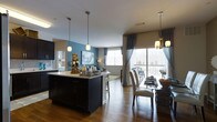 This single-floor residence showcases a thoughtfully designed open-concept layout that seamlessly connects the kitchen, dining, and living areas into one cohesive and inviting space. The kitchen is anchored by a striking dark-cabinetry island with a light countertop, above which pendant lights hang to create a warm, focused ambiance. The bold teal accent wall adds a vibrant pop of color that energizes the space and complements the neutral tones throughout. Rich hardwood flooring runs continuously across the open floor plan, lending warmth and visual continuity. The dining area is elegantly appointed with a glass-top table and upholstered chairs, framed by floor-to-ceiling curtains that soften the natural light pouring in through large sliding glass doors. These doors open directly onto a private balcony, extending the living experience outdoors and offering views of the surrounding landscape. The living room beyond benefits from generous natural light through additional windows, creating a bright and airy atmosphere. The combination of modern finishes, considered lighting, and an open layout makes this space both functional and refined, ideal for everyday living and entertaining alike.