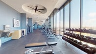 This expansive fitness center occupies a single floor high above the city, offering a well-appointed and thoughtfully designed workout environment. Floor-to-ceiling glass windows line the perimeter, flooding the space with natural light and framing sweeping panoramic views of the surrounding cityscape—an inspiring backdrop for any workout.

The facility is organized into distinct functional zones. The cardio area features a generous row of Life Fitness treadmills, elliptical trainers, and recumbent and upright stationary bikes, all positioned to take full advantage of the window views. Large-format tile flooring throughout provides a clean, durable surface underfoot.

The strength training section includes a comprehensive arrangement of free weights, dumbbells on tiered racks, adjustable benches, and a power rack with barbell and plate storage. Cable machines and additional strength equipment are positioned along the interior walls, accommodating a wide variety of training styles.

Architectural details include oval ceiling recesses fitted with oversized ceiling fans, recessed lighting, and full-length mirrors that visually expand the space. A separate enclosed area with glass-panel doors appears to offer private training or studio space. The overall design is sleek and contemporary, with a cool blue-gray color palette that contributes to a calm, focused atmosphere.