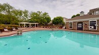 Located at Harpers Crossing in Langhorne, Pennsylvania, this outdoor amenity area is centered around a generously sized, freeform swimming pool with clear turquoise water and a classic brick-edged surround. The pool deck offers ample space for relaxation, furnished with multiple chaise lounge chairs and shaded umbrella tables arranged throughout the patio. A separate, shallower wading pool sits adjacent to the main pool, providing a dedicated area for leisure or younger guests, complete with its own lounge seating and iron safety fencing.

A white pergola structure anchors one end of the outdoor space, offering a shaded gathering area framed by mature trees and well-maintained landscaping. A stone-accented clubhouse building with a chimney is positioned nearby, providing an architectural focal point and additional amenity access for residents. Retaining walls of natural stone define the perimeter of the pool area, adding a refined, cohesive aesthetic to the outdoor environment.

The surrounding residential buildings, constructed in a traditional colonial architectural style with neutral siding and large windows, are visible from the pool area and contribute to the enclosed, community-oriented feel of the space. Mature trees throughout the grounds provide natural shade and privacy, enhancing the overall ambiance of this well-appointed outdoor retreat.