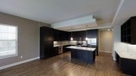 This single-floor space showcases a contemporary design aesthetic centered around a well-appointed, open-concept kitchen. The kitchen is a standout feature, equipped with rich dark espresso cabinetry that extends to the ceiling, providing generous storage throughout. A central island with a light granite countertop serves as both a prep space and a casual gathering point, complemented by modern pendant lighting that hangs above at varying heights, adding a stylish, layered lighting effect. Stainless steel appliances — including a refrigerator, range, and microwave — bring a polished, professional feel to the cooking space. A subway tile backsplash runs along the walls, adding subtle texture and visual interest between the upper and lower cabinetry. The warm hardwood flooring flows seamlessly throughout the space, grounding the dark tones of the kitchen and creating a cohesive, inviting atmosphere. A tray ceiling detail above the kitchen zone adds architectural depth and definition to the open layout. The overall ambiance is modern and sophisticated, with a carefully balanced contrast between dark cabinetry, light countertops, and warm wood tones that gives the space a timeless, upscale character.
