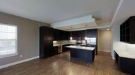 This single-floor space showcases a contemporary design aesthetic centered around a well-appointed, open-concept kitchen. The kitchen is a standout feature, equipped with rich dark espresso cabinetry that extends to the ceiling, providing generous storage throughout. A central island with a light granite countertop serves as both a prep space and a casual gathering point, complemented by modern pendant lighting that hangs above at varying heights, adding a stylish, layered lighting effect. Stainless steel appliances — including a refrigerator, range, and microwave — bring a polished, professional feel to the cooking space. A subway tile backsplash runs along the walls, adding subtle texture and visual interest between the upper and lower cabinetry. The warm hardwood flooring flows seamlessly throughout the space, grounding the dark tones of the kitchen and creating a cohesive, inviting atmosphere. A tray ceiling detail above the kitchen zone adds architectural depth and definition to the open layout. The overall ambiance is modern and sophisticated, with a carefully balanced contrast between dark cabinetry, light countertops, and warm wood tones that gives the space a timeless, upscale character.