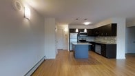 This single-floor space offers a thoughtfully designed open-concept layout that seamlessly connects the kitchen and living areas, creating a bright and functional environment. The kitchen is a standout feature, showcasing dark espresso cabinetry that contrasts beautifully with a light stone-topped center island, providing both additional prep space and a casual gathering spot. Pendant lighting hangs above the island, adding a modern, decorative touch, while flush ceiling fixtures ensure the space is well-lit throughout. A full suite of stainless steel appliances — including a refrigerator, range, microwave, and dishwasher — lines the kitchen walls, complemented by a mosaic tile backsplash that adds texture and visual interest. Warm hardwood-style flooring runs throughout the space, contributing to a cohesive and inviting ambiance. The open living area benefits from the flow of the layout, offering ample room for furnishing and personalization. A baseboard heating system runs along the perimeter, ensuring year-round comfort. A private entry door provides direct access, and the clean, neutral wall tones create a versatile backdrop suited to a variety of interior styles. Overall, this space combines practical functionality with contemporary design details.