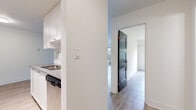 This thoughtfully laid-out one-bedroom, one-bathroom unit offers a clean, modern aesthetic with light-toned laminate flooring and crisp white walls throughout. The galley-style kitchen is a functional highlight, featuring white shaker cabinetry, white subway tile backsplash, grey quartz countertops, a stainless steel sink, and a full suite of stainless steel appliances including a refrigerator, electric range, built-in microwave, and dishwasher. A walk-in closet or storage area is accessible directly from the kitchen corridor, adding practical convenience. A bold black three-panel interior door adds a striking contrast to the otherwise neutral palette.

The living room is bright and inviting, anchored by a full-width sliding glass door that opens to a private patio area surrounded by lush greenery, bringing natural light and outdoor views into the space. Baseboard heating runs along the perimeter walls. The bedroom is comfortably sized, featuring a large window overlooking a green outdoor area, laminate flooring, and a baseboard heater.

The bathroom is finished to a high standard, with floor-to-ceiling white subway tile surrounding the soaker tub and shower, grey slate-style floor tile, a white vanity with an undermount sink, and a mirrored medicine cabinet.