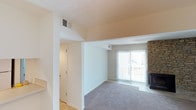 This single-floor residence offers a clean, functional layout with a welcoming living area at its heart. The living room is finished with plush wall-to-wall carpeting in a neutral gray tone, providing a comfortable and versatile foundation for the space. A standout architectural feature is the brick fireplace positioned along one wall, adding a classic, cozy character to the room. Natural light flows generously into the living area through a large sliding glass door dressed with vertical blinds, which opens onto an outdoor balcony — offering both ventilation and an exterior connection. The walls are finished in crisp white and warm neutral tones, contributing to a bright and airy atmosphere throughout. Ceiling-mounted HVAC vents are integrated cleanly into the design, ensuring year-round climate comfort. A transitional hallway connects the living area to additional rooms, with a closed door visible along the corridor suggesting private spaces beyond. The entryway area features hard flooring, providing a practical transition zone before giving way to the carpeted main living space. Overall, the layout is straightforward and efficient, with architectural details like the brick fireplace and sliding glass door adding warmth and character to the interior.