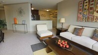 This single-floor residence showcases an open-concept design that seamlessly integrates the living area with the kitchen. The living space is furnished with clean-lined, modern seating — including armless chairs and a sofa with neutral upholstery complemented by warm wood frames — arranged around a dark wood coffee table. A decorative bowl of red ornamental fruit serves as a focal centerpiece. The space is grounded by a mix of carpet and area rugs, adding texture and definition to the layout. A side table with a warm-toned lamp provides ambient lighting alongside the ceiling-mounted fixtures. The kitchen, visible beyond a half-wall partition, features dark cabinetry, a stainless steel refrigerator, and a light-colored countertop that doubles as a breakfast bar. Decorative wall art, including a sculptural swan motif and a series of horizontally arranged accent pieces, adds visual interest throughout. Neutral gray walls and consistent flooring unify the space, giving it a calm, contemporary ambiance throughout.