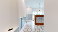 Located at 6170 Kitt Ln NW in Huntsville, Alabama, this mail and package room serves as a functional yet thoughtfully designed communal space within The Kelvin community. The hallway layout flows into a more open mail center area, unified by striking geometric-patterned tile flooring in neutral gray and white tones that adds visual interest throughout the space. Crisp white walls and high ceilings create a bright, open atmosphere, enhanced by abundant natural light streaming through large windows fitted with roller shades. A prominent central island clad in warm wood houses an array of individual mailboxes, providing residents with convenient mail access. Additional built-in mailbox units and secure package lockers line the surrounding walls, ensuring comprehensive mail and delivery management. Overhead, a series of industrial-style pendant lights with cage and globe fixtures add a warm, modern-vintage character to the space. A connecting hallway leads to adjacent community areas, offering glimpses of wood-toned ceilings and light hardwood flooring beyond. The bold gold lettering of "THE KELVIN" displayed on the wall makes for a striking, branded focal point that anchors the room's identity and reinforces the community's polished, contemporary aesthetic.