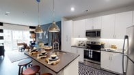 This single-floor space showcases a thoughtfully designed open-concept layout that seamlessly connects the kitchen and living areas. The kitchen is a standout feature, boasting crisp white cabinetry that extends to the ceiling, offering generous storage throughout. Dark countertops provide a striking contrast against the white cabinetry, while a textured geometric tile backsplash adds visual depth and character to the space.

A central island with matching dark countertops serves as both a prep area and casual dining spot, accommodated by bar-style seating. Two polished metallic pendant lights hang above the island, delivering focused task lighting while contributing a refined, industrial-chic aesthetic. Stainless steel appliances, including a range and over-the-range microwave, complete the kitchen's modern appeal.

The adjoining living area is visible beyond the kitchen, anchored by a bold teal-blue accent wall adorned with framed artwork. The open flow between spaces creates a sense of expansiveness, while the cohesive color palette of navy, white, and metallic tones ties the entire area together into a contemporary, well-styled interior.