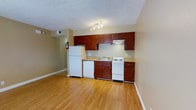 This single-floor space offers a practical and well-organized layout designed for everyday comfort and functionality. The kitchen serves as a central feature, showcasing rich, dark reddish-brown cabinetry that provides ample upper and lower storage. The countertops offer a clean, light work surface, complemented by a double-basin sink and a full suite of white appliances, including a refrigerator, dishwasher, electric range, and a range hood above the cooktop. A track light fixture overhead ensures the kitchen area is well-lit for meal preparation. The warm, light-toned hardwood flooring extends throughout the space, adding a sense of warmth and continuity to the interior. Neutral beige walls keep the atmosphere bright and open, while clean white baseboards provide a crisp finish. The textured ceiling adds a classic touch to the overall aesthetic. A fire safety extinguisher is conveniently mounted near the refrigerator, reflecting a practical approach to the space's design. Visible HVAC venting integrated into the walls ensures year-round climate comfort. Overall, the space presents a clean, move-in-ready environment with a straightforward, functional character.