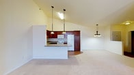 This single-story space showcases an open-concept design that integrates the kitchen and living area into one cohesive, airy environment. The vaulted ceilings rise dramatically, amplifying the sense of spaciousness and contributing to the bright, welcoming atmosphere throughout. Pale yellow walls complement the neutral tones of the space, enhancing the light and open feel.

The kitchen is appointed with rich, dark cherry wood cabinetry that provides ample storage. White appliances, including a refrigerator, range, and built-in microwave, are neatly integrated into the cabinetry layout. A peninsula with a light-toned countertop extends into the living space, creating a natural divide while maintaining visual connectivity. Three pendant lights with stained glass shades hang above the peninsula, adding a decorative touch and functional illumination.

The adjoining living area features soft, plush carpeting in a warm neutral tone, offering a comfortable contrast to the hardwood flooring visible in the kitchen. An additional pendant light fixture with a stained glass design echoes the kitchen's style, tying the two spaces together cohesively. The overall ambiance is clean, functional, and inviting.