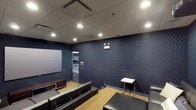 This single-floor screening room is thoughtfully designed to deliver a focused and immersive viewing or presentation experience. The space is defined by its striking deep navy blue walls adorned with a bold chevron pattern, lending a dynamic and sophisticated visual character to the interior. A large projection screen anchors one end of the room, making it well-suited for film screenings, presentations, or media events. Recessed lighting embedded in a sleek drop ceiling provides controlled, even illumination throughout, while wall-mounted sconce fixtures add a refined touch and contribute to the room's theater-like ambiance. Rows of dark leather seating are arranged in a tiered layout, ensuring clear sightlines to the screen from every seat. A small side table is conveniently positioned near the seating area for added functionality. The flooring features a warm-toned carpet that complements the darker tones of the walls and furnishings, enhancing the room's acoustics and comfort. A doorway at the rear provides access to an adjacent area visible through the opening. Overall, the space strikes a balance between professional utility and cinematic atmosphere, making it a versatile venue for both business and entertainment purposes.