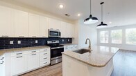 This well-designed single-floor unit showcases a thoughtful open-concept layout that seamlessly connects the kitchen and living areas. The kitchen is a standout feature, boasting crisp white shaker cabinetry with matte black hardware, granite countertops, and a striking navy blue subway tile backsplash. A central island with an undermount sink and a brushed gold faucet provides additional prep space, while stylish industrial-inspired pendant lights hang overhead. Stainless steel appliances — including a refrigerator, electric range, microwave, and dishwasher — complete the modern culinary space. Warm-toned luxury vinyl plank flooring flows throughout the main living areas, lending a cohesive and inviting feel. The open living area benefits from abundant natural light through a bank of windows that offer views of the surrounding exterior. The bedroom features plush carpeting and a large window with outdoor views, creating a comfortable and tranquil retreat. The bathroom is clean and functional, featuring wood-look LVP flooring consistent with the rest of the unit, a vanity with granite countertops, and a tiled shower area. The overall aesthetic is modern and move-in ready, with a neutral color palette and quality finishes throughout.
