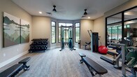 Located at 1302 Rio Valley Dr in Raleigh, NC, this well-appointed fitness center offers a comprehensive workout environment across a single, thoughtfully designed floor. The space is divided into distinct functional zones, each catering to different aspects of physical fitness.

The cardio area is anchored by a row of elliptical trainers and treadmills positioned in front of large floor-to-ceiling windows, allowing natural light to flood the space and providing refreshing outdoor views of the surrounding greenery. A ceiling fan overhead and bold geometric green and neutral wall murals give this zone a vibrant, energetic atmosphere.

A dedicated strength training room features a cable machine, adjustable bench, dumbbell rack, and additional resistance equipment, all set within a bright bay-windowed alcove that maximizes natural light and openness.

The cycling studio is equipped with multiple stationary bikes, stability balls, and a large wall-mounted flat-screen TV, complemented by full-length mirrors and hardwood-style flooring, creating an immersive group fitness setting.

Throughout the facility, a striking chevron-patterned tile floor anchors the central corridor, which also houses a dual water fountain station and clearly marked restroom facilities. Decorative laser-cut green panels add a distinctive architectural flair. Recessed lighting and ceiling fans ensure a comfortable environment, completing this modern, amenity-rich fitness center.