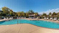 Located at 3325 Oak Forest Dr in Fayetteville, North Carolina, this outdoor amenity area centers around a generously sized freeform swimming pool with a distinctive curved layout that offers both shallow and deeper swimming zones. The pool's crystal-clear water is complemented by a wide, smooth concrete sundeck that surrounds it on all sides, providing ample space for lounging and recreation. Numerous chaise lounge chairs and seating areas are arranged throughout the deck, many shaded by navy blue and teal umbrellas for comfort on sunny days. A white pergola structure anchors one end of the pool area, offering additional shaded seating with casual outdoor furniture beneath it. The space is bordered by a black iron fence that encloses the area while maintaining open sightlines to the surrounding mature trees, including tall pines and palm trees that add a lush, resort-like ambiance. A brick clubhouse building with a pitched roof sits adjacent to the pool, providing a functional anchor to the space. The entire area benefits from abundant natural light and open sky views, creating a bright and inviting atmosphere ideal for outdoor leisure and relaxation.