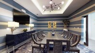 This single-floor space is a well-appointed meeting or private dining room designed with a bold, contemporary aesthetic. The walls are finished in striking horizontal blue and white stripes, creating a dynamic visual rhythm that defines the room's character. A tray ceiling with integrated ambient lighting adds architectural depth and a refined sense of elevation to the space.

At the center of the room sits a substantial rectangular table crafted from dark, textured wood, surrounded by a set of curved, upholstered chairs in a charcoal finish that blend style with comfort. Overhead, a sculptural sputnik-style chandelier with globe bulbs serves as a dramatic focal point, complementing the room's modern sensibility.

Along one wall, a dark navy sideboard provides functional storage while reinforcing the room's cohesive color palette. A large flat-screen television is mounted nearby, making the space well-suited for presentations or media-driven gatherings. Decorative gold artwork panels and tall dried botanicals in a white vase add artistic touches, while a patterned area rug grounds the seating arrangement. The overall ambiance is sophisticated, bold, and versatile.