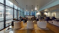 Located in the heart of Chicago's vibrant River North neighborhood, this single-floor amenity lounge at 668 N La Salle Dr offers a sophisticated and well-appointed communal space designed for comfort and socializing. The main lounge area is anchored by floor-to-ceiling windows that span the length of the room, flooding the space with natural light and providing sweeping views of the surrounding Chicago skyline and cityscape. Pendant lighting with sleek, cone-shaped fixtures hangs from the ceiling, complementing the modern aesthetic throughout. The lounge is thoughtfully arranged with a variety of seating options, including plush leather sofas, rounded upholstered chairs, and accent armchairs grouped around coffee tables atop patterned area rugs, creating intimate conversation zones. Polished concrete floors run throughout, lending an industrial-chic character to the space. An adjacent lounge area features a large sectional sofa and a gallery wall of black-and-white Chicago photography, adding a locally inspired artistic touch. A compact kitchenette is equipped with dark cabinetry, white countertops, a stainless steel refrigerator, and open shelving with decorative accents, making the space fully functional for gatherings. A game table and additional seating areas round out this versatile, amenity-rich environment.