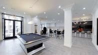 This single-floor clubhouse is thoughtfully designed to offer a refined and versatile social environment. The space is anchored by a striking monochromatic color palette — crisp white walls, light gray tile flooring, and bold black accents — creating a clean, contemporary aesthetic throughout. Decorative white columns with crown molding details add a touch of architectural elegance, complementing the recessed lighting that bathes the entire space in a bright, welcoming glow. A standout feature is the full-size billiards table, positioned prominently in the game area and illuminated by a modern pendant light fixture, making it an immediate focal point. Adjacent to the game area, a lounge and bar section offers high-top seating with sleek black barstools arranged around tall tables, ideal for socializing or casual gatherings. A dramatic egg-shaped accent chair adds a sculptural design element to the seating area. Toward the back, a darker, intimate bar counter with bar seating provides an additional gathering spot. The open layout encourages flow between activity zones, while large windows allow natural light to filter in. The overall ambiance is upscale, modern, and inviting — well-suited for community events and leisure activities alike.